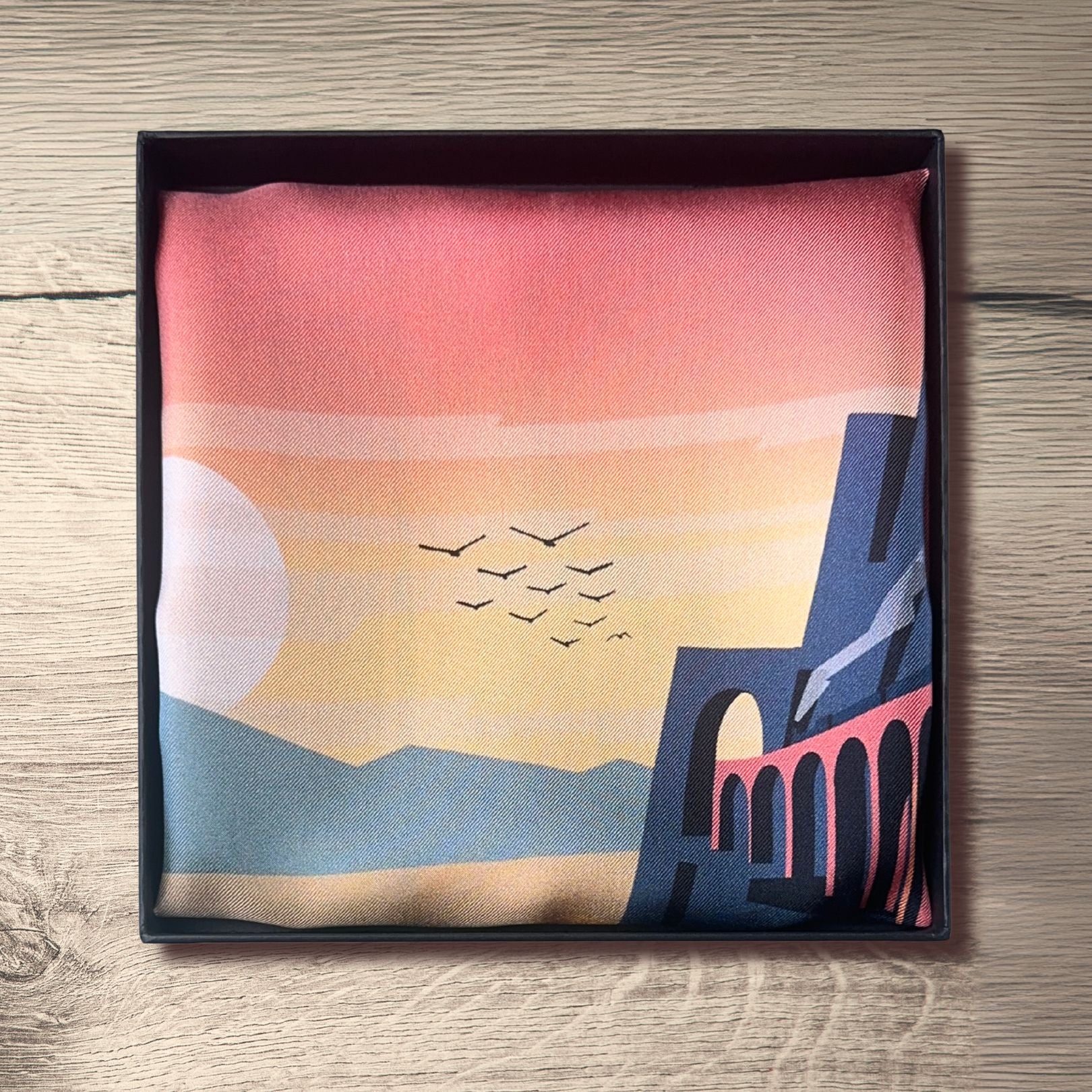 L'Odysee silk Pocket Square with a scenic design of a bridge and birds, folded in a BLUEMATERIALS box on a wooden surface.