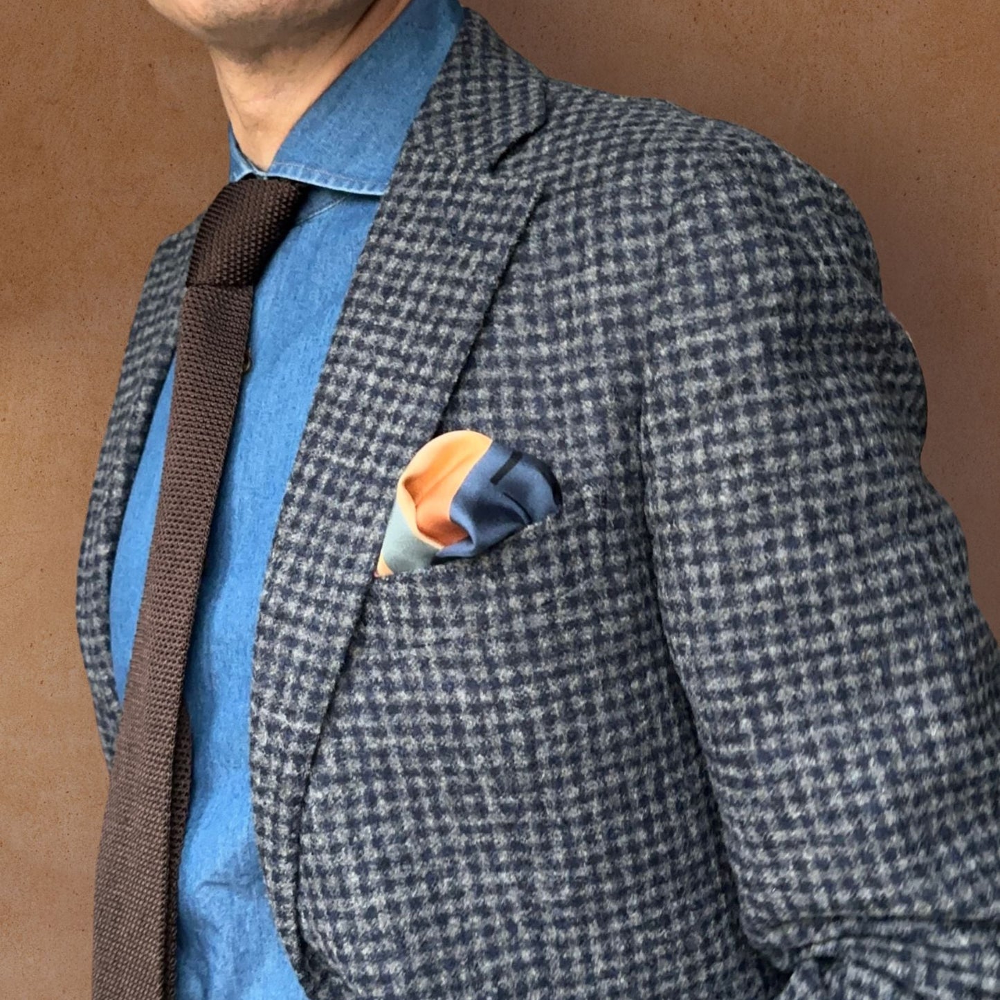 Blue-grey checkered blazer with L'Odyssée, the Amalfi inspired pocket square and blue denim shirt on a brown background