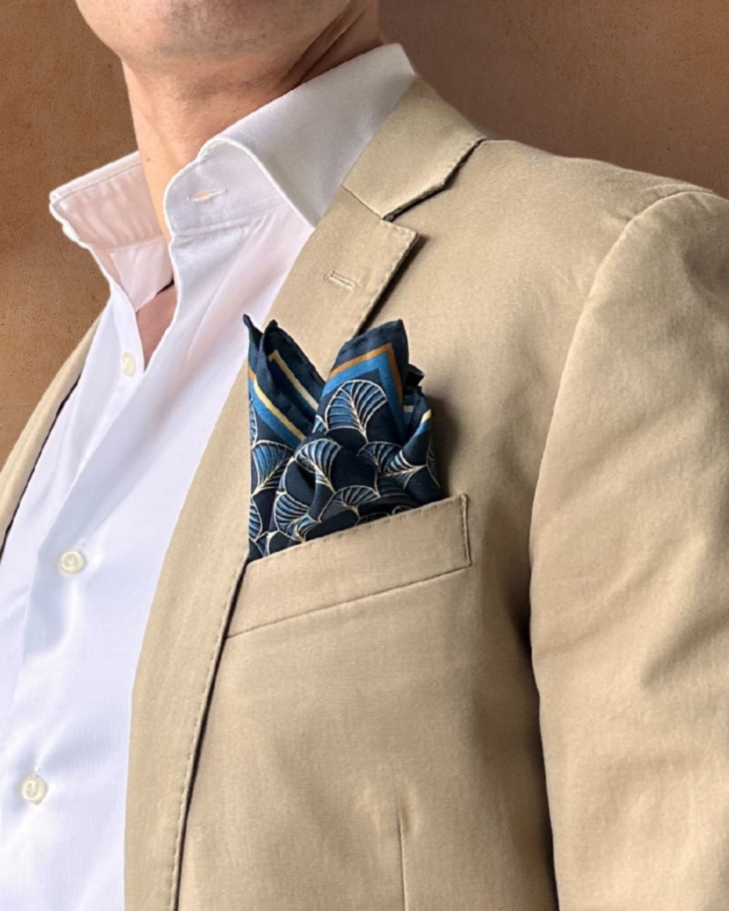 Midnight blue and gold pocket square in a lapel pocket of a cream jacket paired with a white shirt on a tobacco background