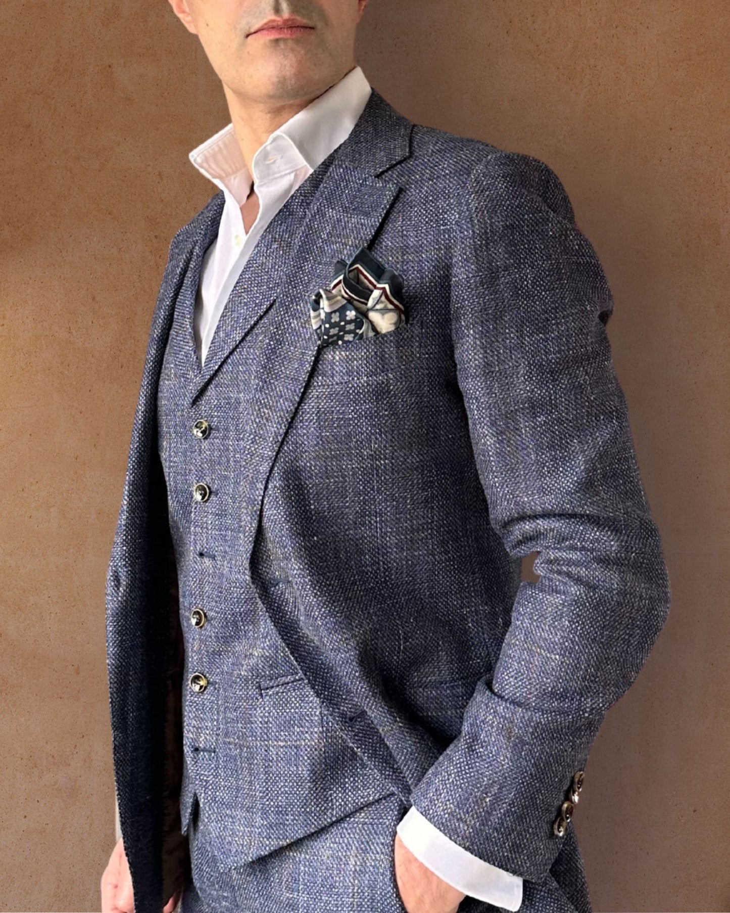Blue cream and burgundy pocket square on the lapel pocket of a blue-grey textured suit paired with a crisp white shirt on a tobacco background