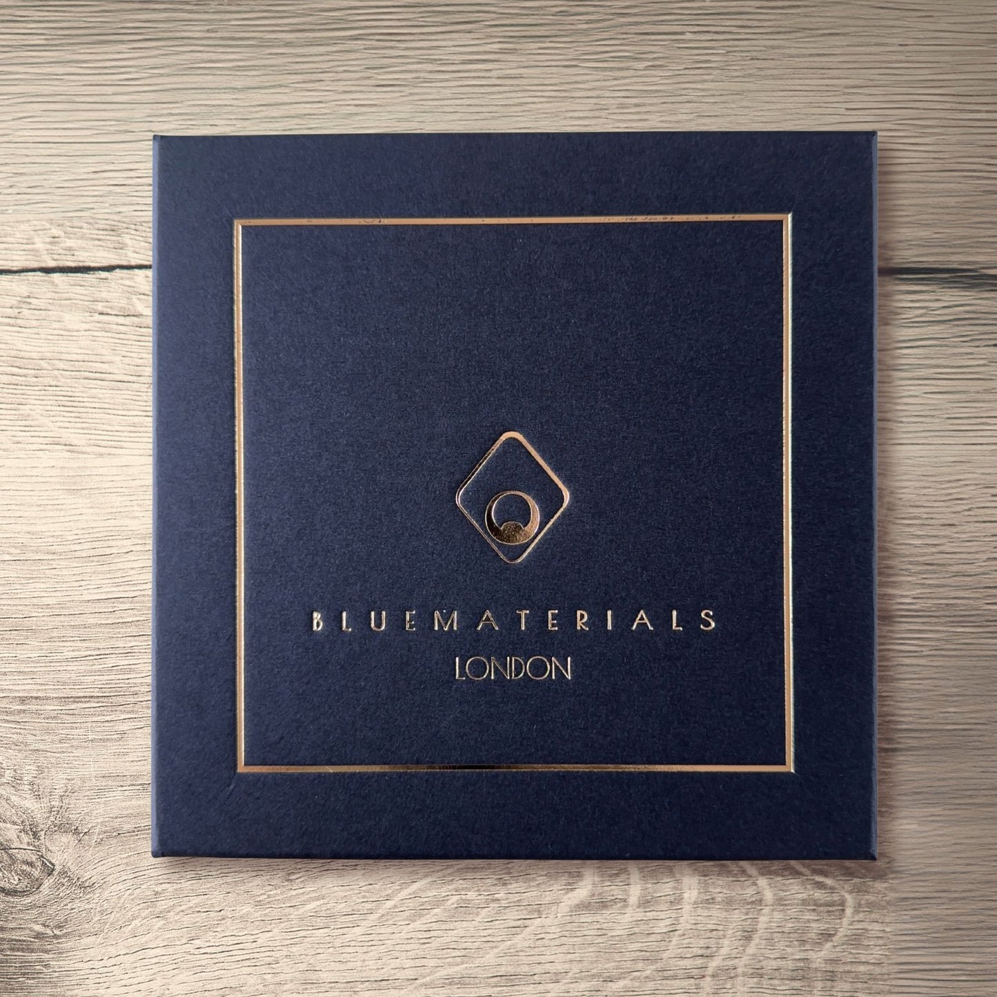BLUEMATERIALS gift box cover with a golden embossed logo and decorative frame, on a rustic wooden table