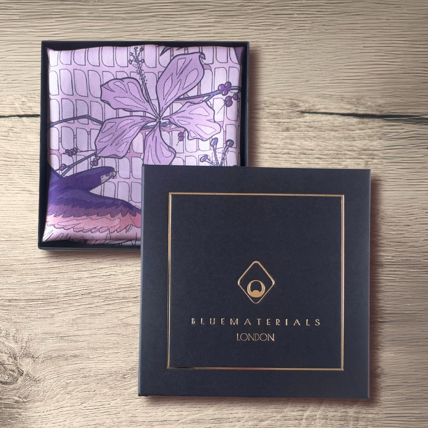 Pink purple, and navy blue pocket square folded inside a gift box next to BLUEMATERIALS cover with a golden embossed logo and decorative frame, on a rustic wooden table