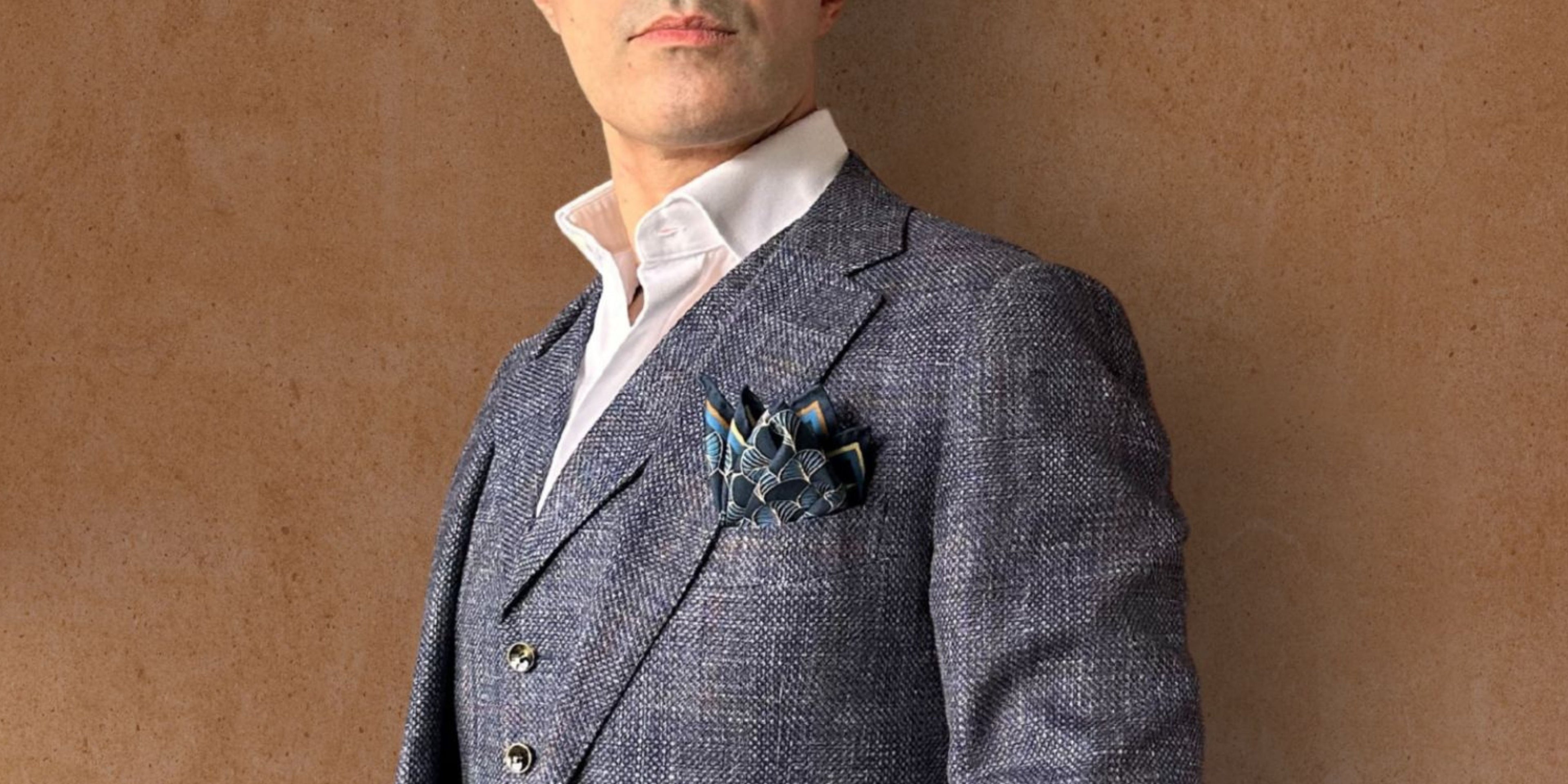 Man wearing a gray suit with a white shirt and blue and gold pocket square against a brown background
