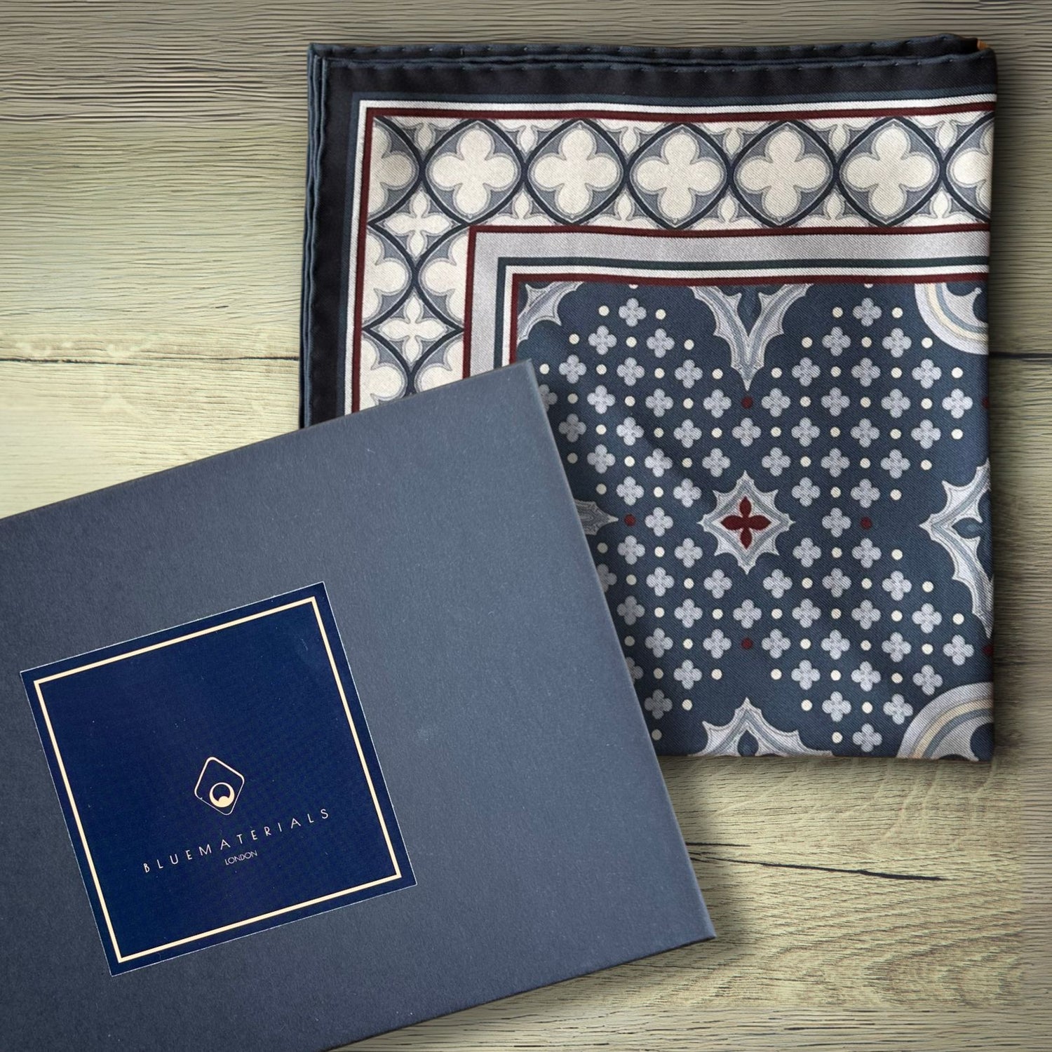 Blue cream and burgundy pocket square folded on a wooden rustic table next to a dark grey gift box with BLUEMATERIALS branding and logo