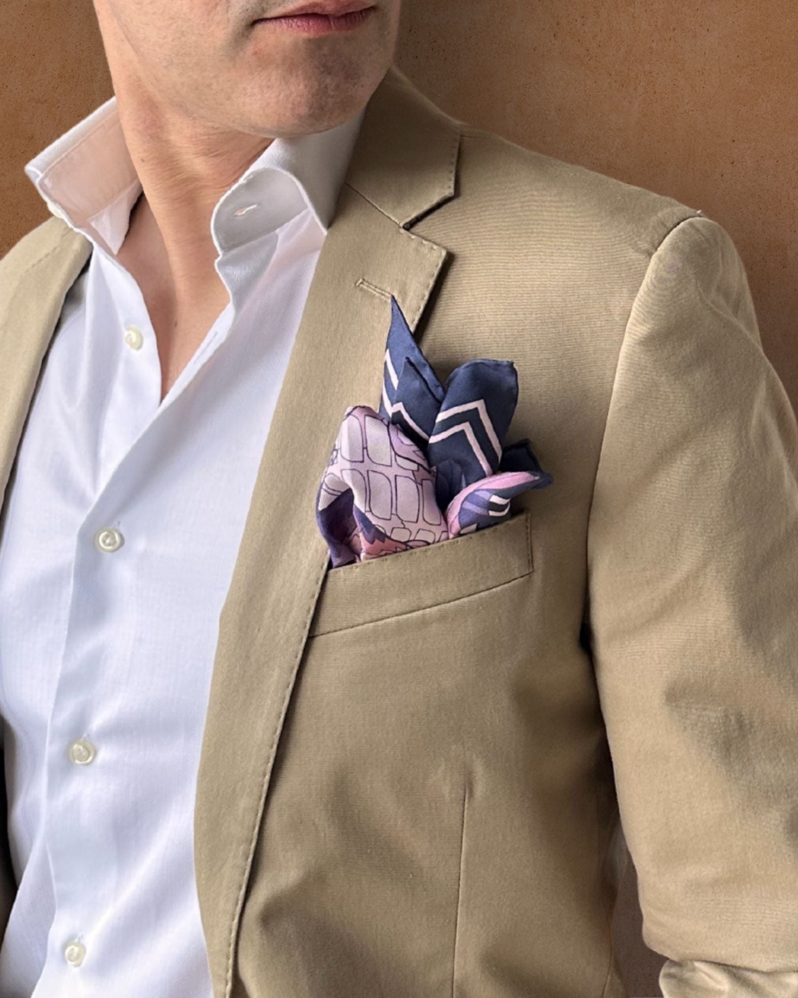 Pink and navy blue pocket square with peak fold on a lapel pocket of a cream jacket paired with a white shirt on a tobacco background