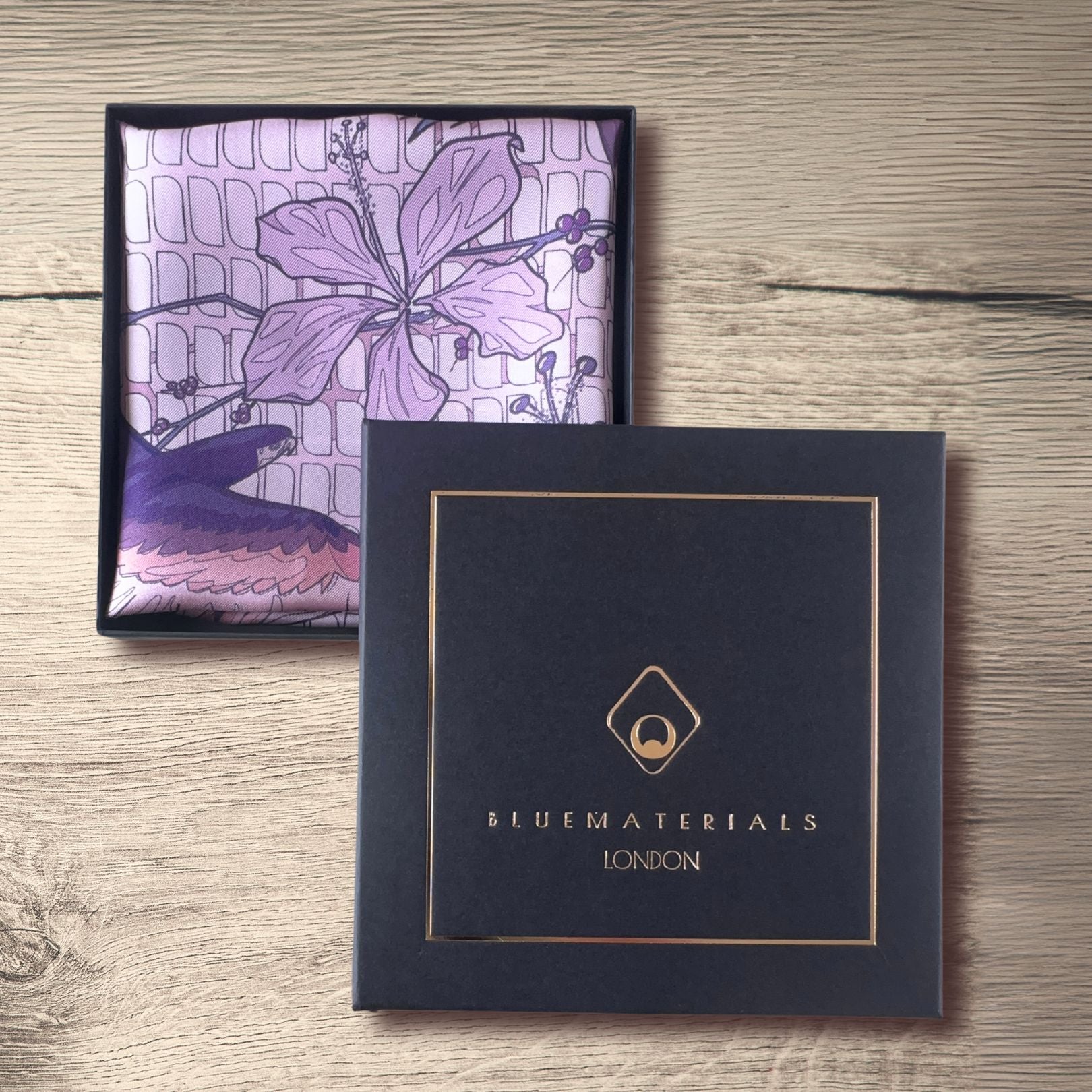 Pink purple, and navy blue pocket square folded inside a gift box next to  BLUEMATERIALS cover with a golden embossed logo and decorative frame, on a rustic wooden table 